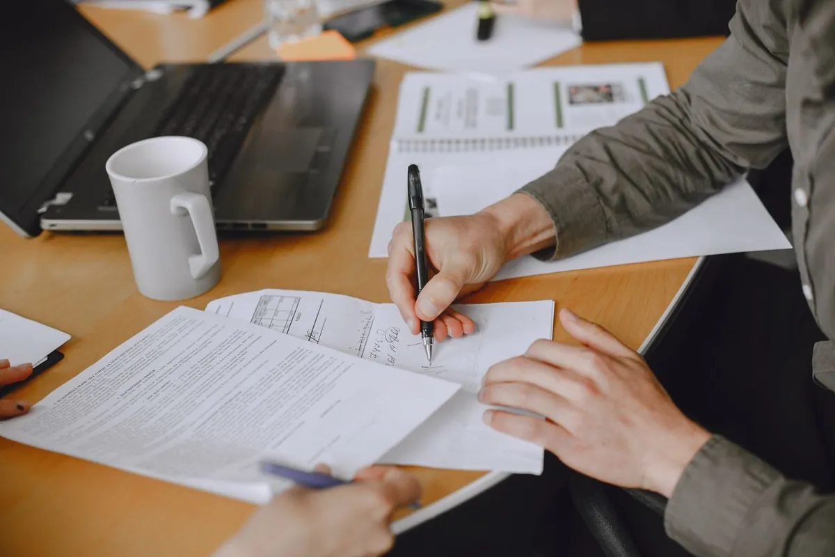 Mãos de uma pessoa assinando um documento com caneta sobre a mesa, com notebook e papéis ao redor