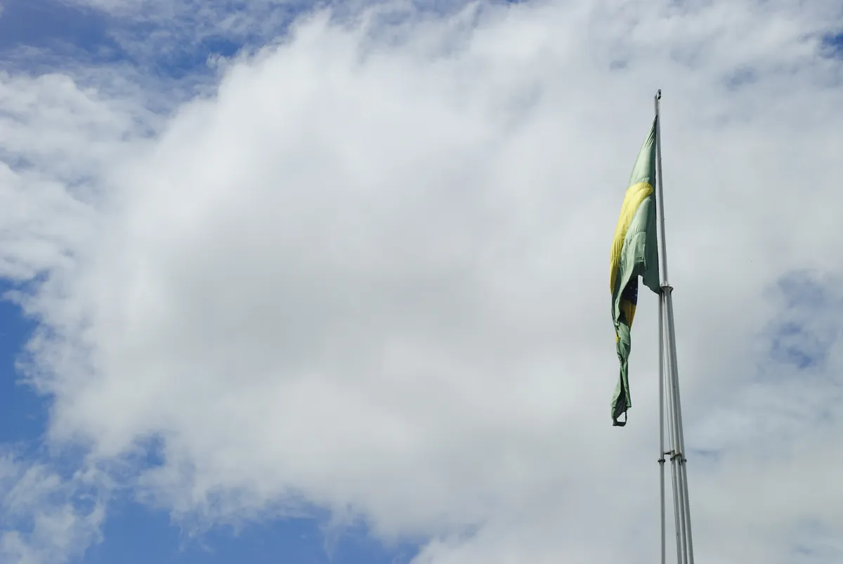 Bandeira do Brasil hasteada em mastro, com o céu nublado ao fundo