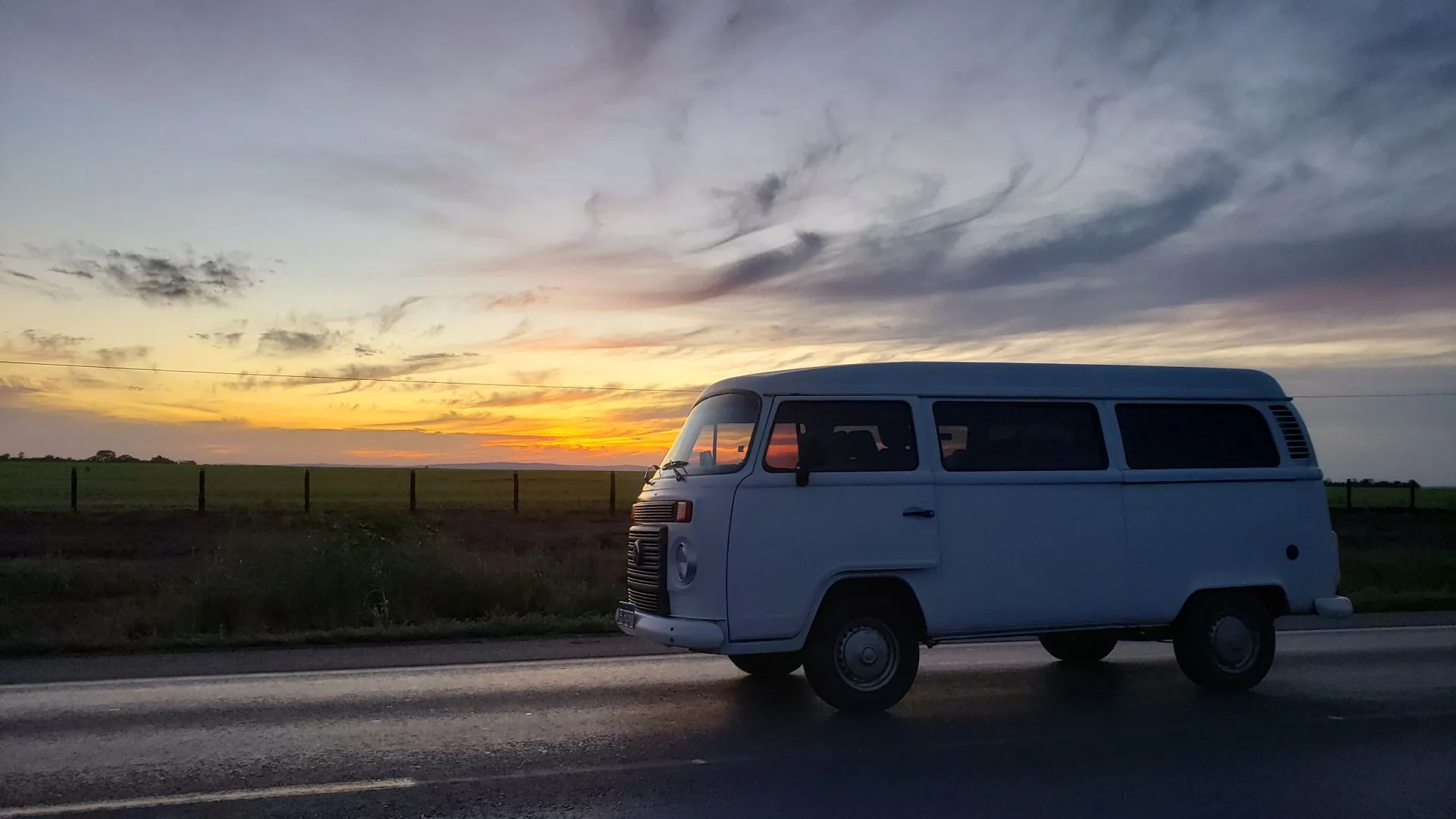 Kombi branca trafegando por uma estrada de Mato Grosso ao pôr do sol, sob um céu amplo e dourado