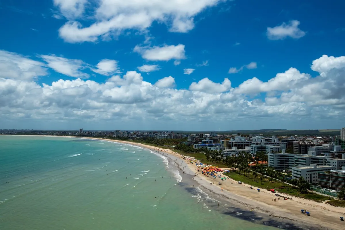 Vista aérea da praia do Cabo Branco, em João Pessoa, com mar verde-azulado, faixa de areia, guarda-sóis coloridos e prédios da orla